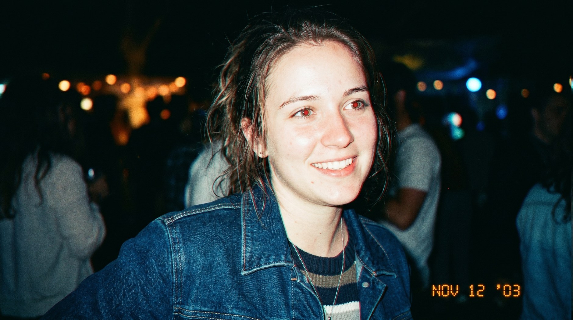 A young woman in denim, lit by direct on-camera flash, with a dated digital timestamp NOV 12 03 in the corner — authentic 2000s digicam aesthetic.